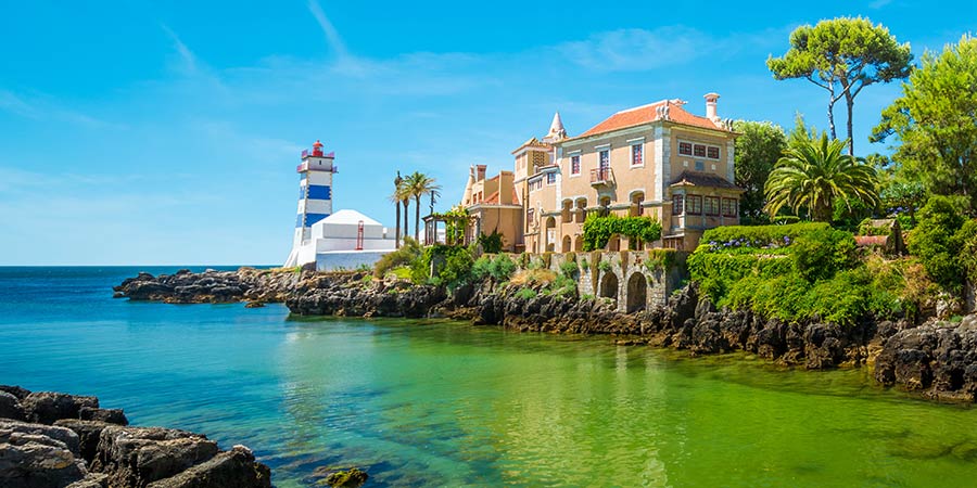 A beautiful stone building stands on a rocky coast next to a white and blue striped lighthouse, both surrounded by lush greenery and emerald sea waters. 